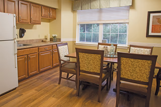 A dining area in a senior living facility with a wooden table surrounded by six chairs featuring patterned upholstery. The room has wooden flooring, a large window with blinds, and a kitchen area with wooden cabinets, a white refrigerator, and a countertop with a sink and decorative items.