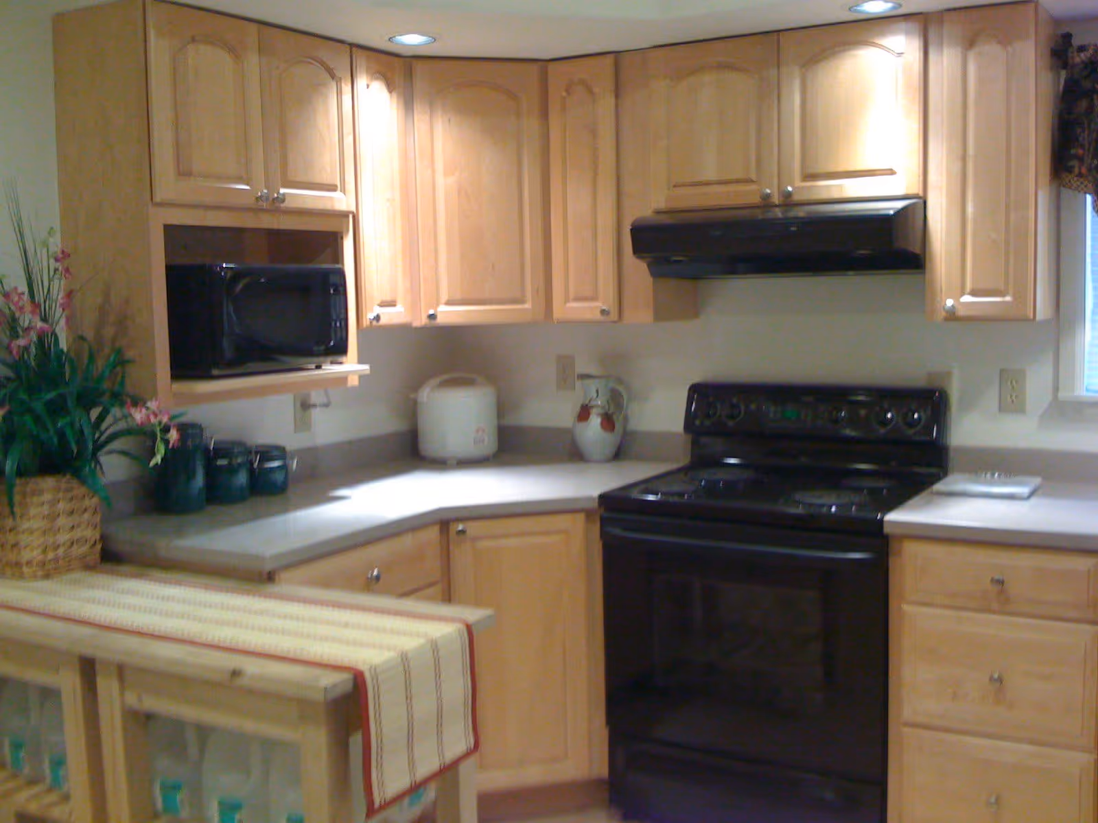A kitchen with light wood cabinets, a black electric stove with oven, a black microwave, a rice cooker, and a decorative pitcher on the countertop. There is a small table or counter with a striped runner and a basket with pink flowers and greenery.