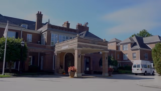 Exterior view of a large brick building with multiple chimneys and windows, featuring a covered entrance with columns and flower pots. A white van is parked on the right side near the building, under a clear blue sky.
