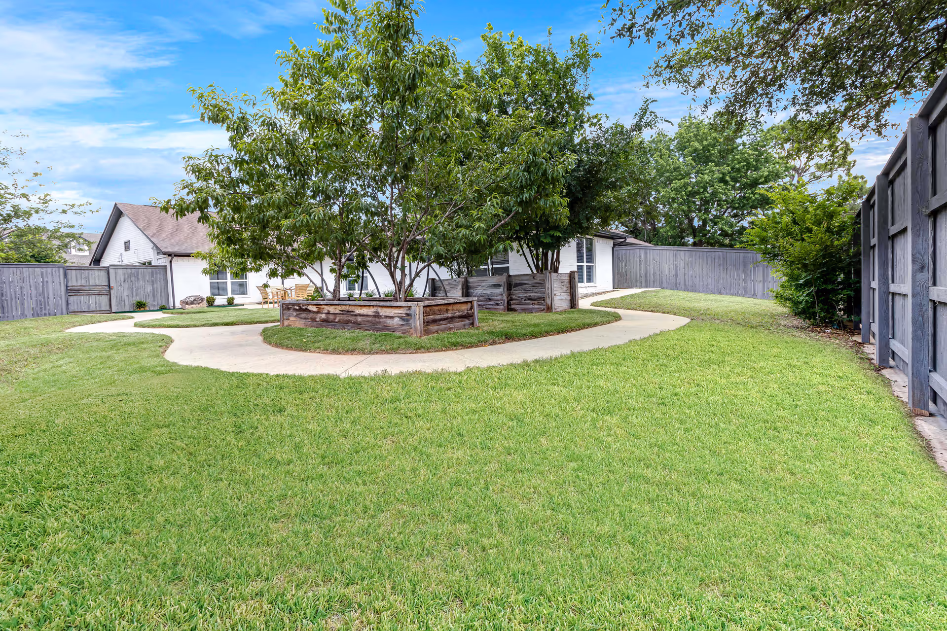A well-maintained outdoor garden area with a curved concrete pathway surrounding a raised wooden planter box filled with green leafy trees. The garden is enclosed by a wooden fence, and a white building with a brown roof is visible in the background under a partly cloudy blue sky.