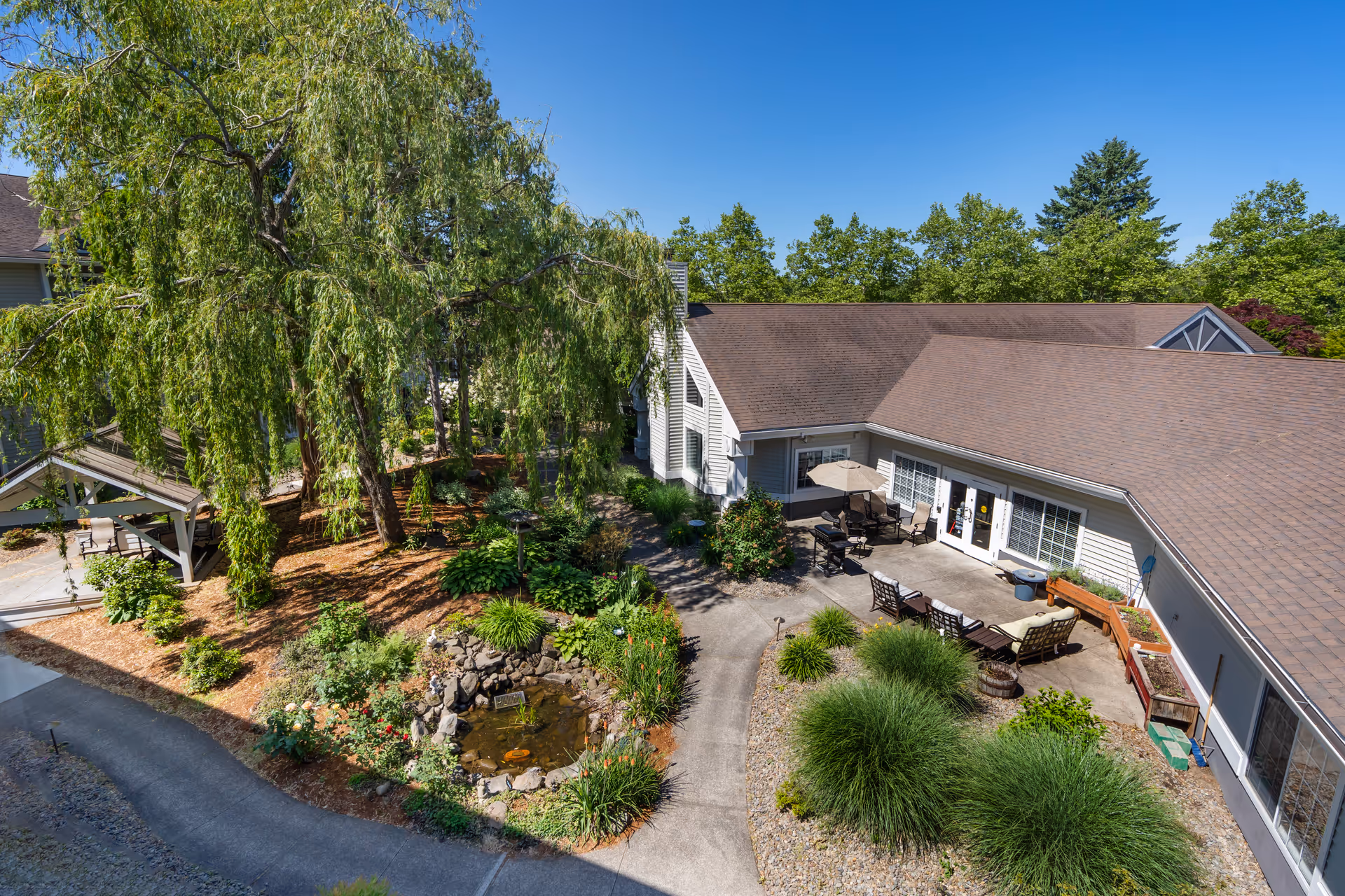 Outdoor courtyard area of a senior living facility with a small pond surrounded by plants and flowers, a large tree providing shade, a covered seating area, and a patio with chairs, tables, and an umbrella next to a building under a clear blue sky.