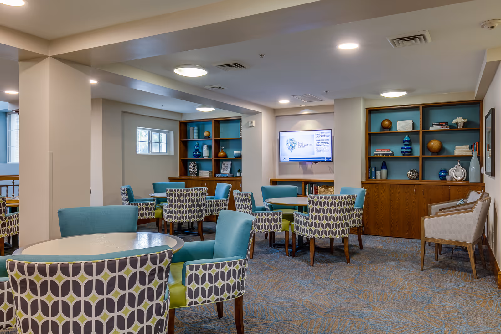 A well-lit common area with multiple round tables surrounded by patterned and solid-colored chairs. The room features built-in wooden shelves with decorative items and books, a wall-mounted television, and carpeted flooring with a blue and beige pattern.