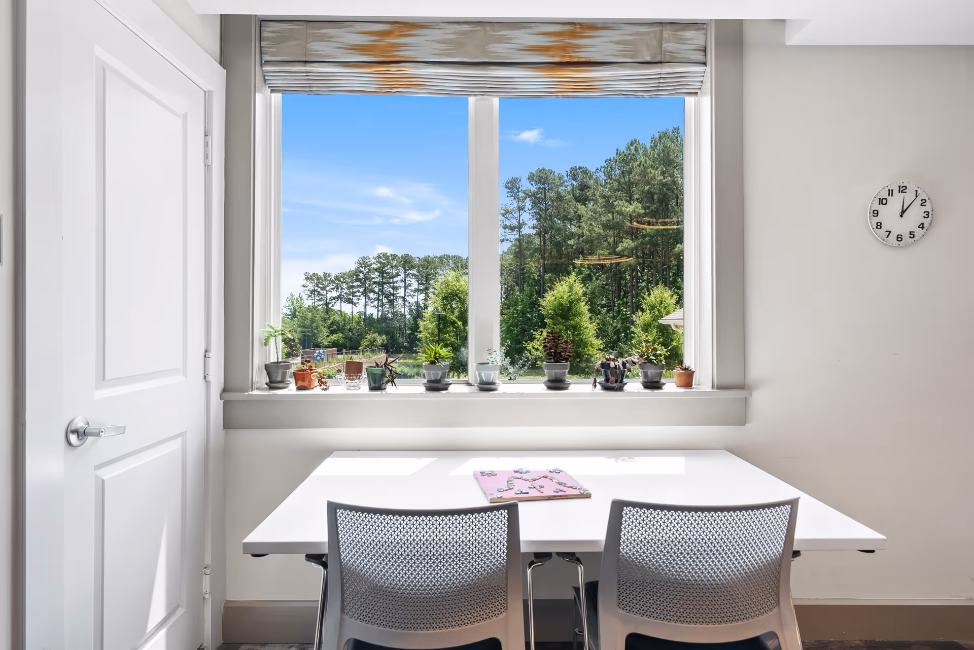 A bright room with a white table and two gray chairs facing a large window. The window sill holds several small potted plants. Outside the window, there is a view of green trees under a clear blue sky. A white door is on the left wall and a round clock showing 12:05 is on the right wall.