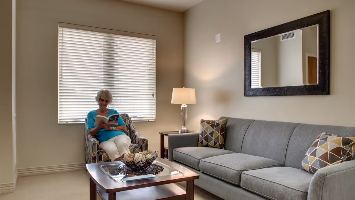 A senior woman sitting in a patterned armchair reading a book in a well-lit living room. The room features a large gray sofa with patterned cushions, a wooden coffee table with decorative items, a side table with a lamp, and a large mirror on the wall.
