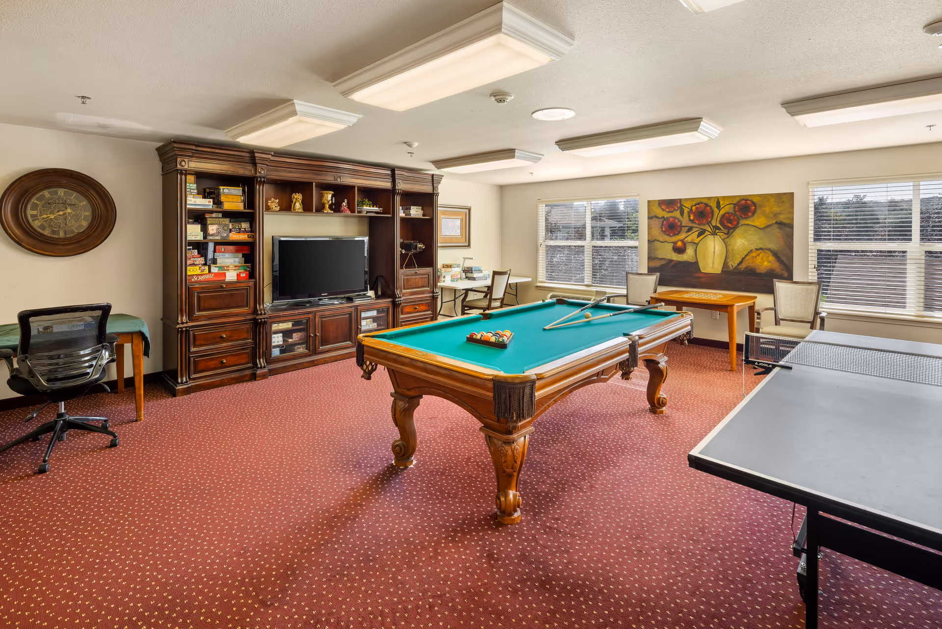 A recreational room with a pool table in the center, a ping pong table on the right, and a large wooden entertainment center with a TV and board games on the left. The room has red carpet with a small pattern, several chairs and tables near windows with blinds, and a large painting of flowers on the far wall.