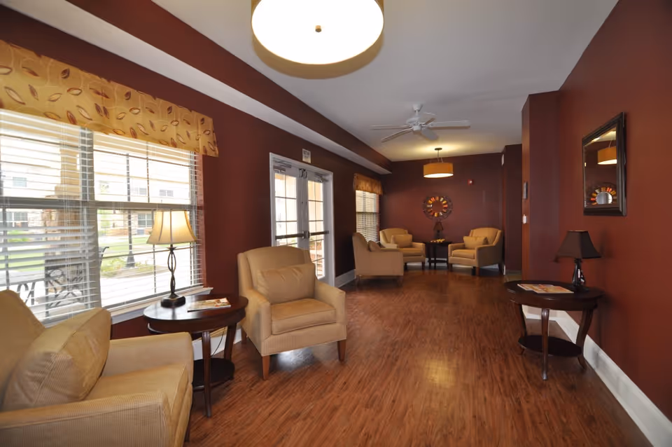 A cozy lounge area with brown walls and wooden flooring featuring several beige armchairs arranged around small round wooden tables. The room has large windows with blinds and patterned valances, ceiling lights, a ceiling fan, and decorative wall mirrors. There are table lamps on the side tables, and the space appears warm and inviting.
