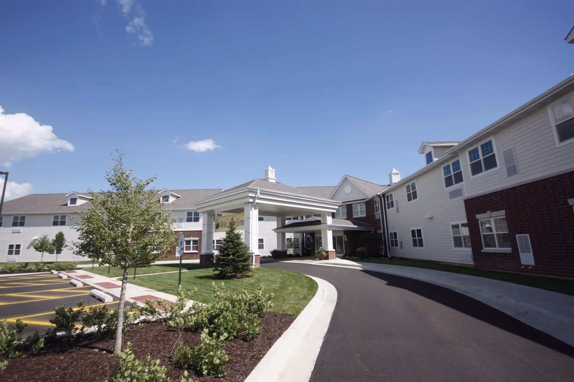 Exterior view of a senior living facility with a covered entrance, landscaped greenery, a curved driveway, and a clear blue sky.