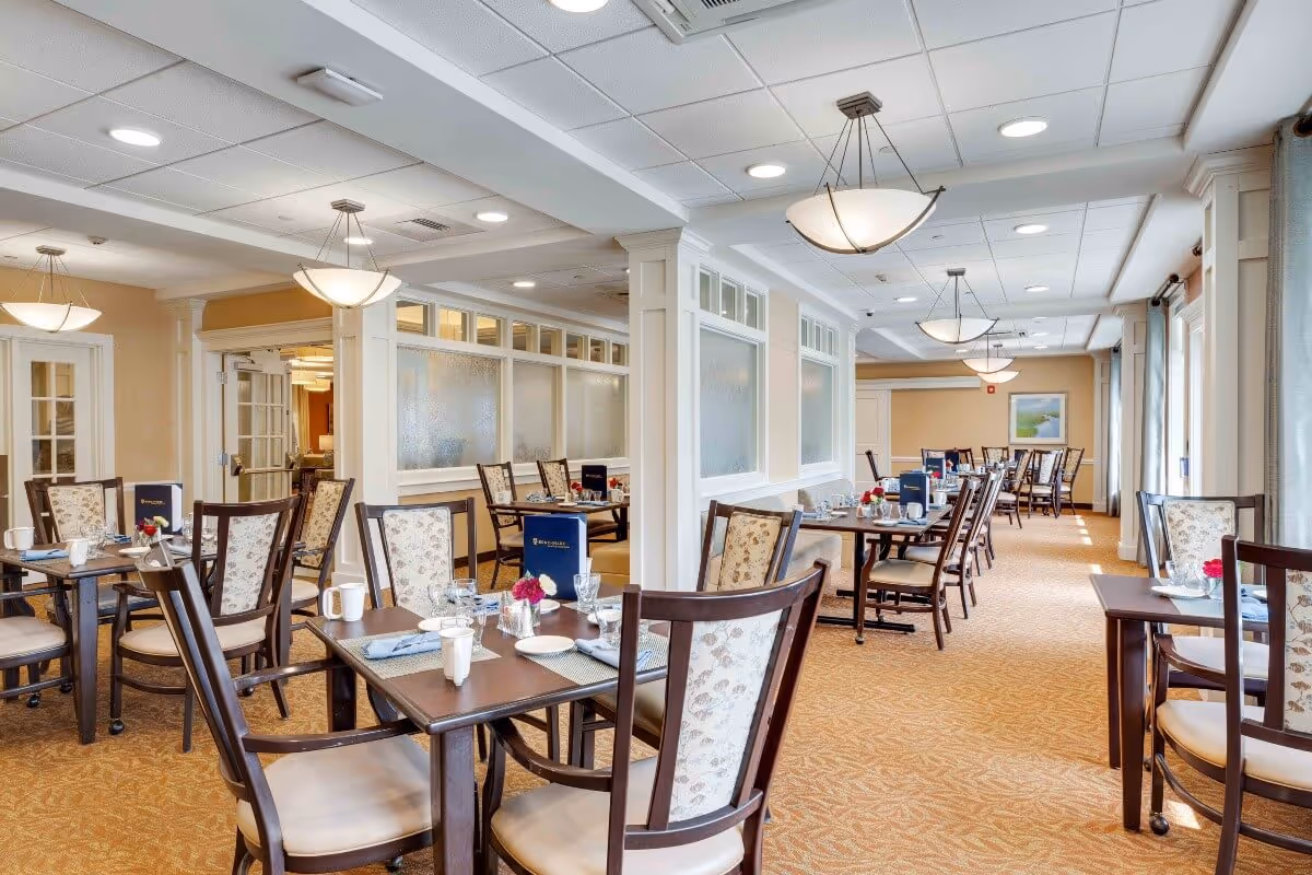 A bright and spacious dining room in a senior living facility with multiple wooden tables and chairs arranged neatly. Each table is set with white cups, glassware, napkins, and small flower vases. The room features beige walls, carpeted flooring, large windows with curtains allowing natural light, and ceiling lights providing additional illumination.