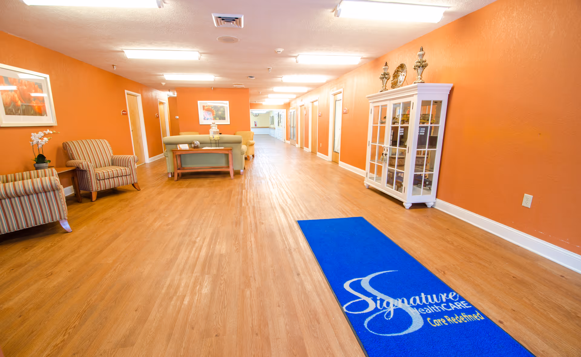 A long hallway in a senior living facility with orange walls and wooden flooring. There are several doors along the hallway, a white display cabinet on the right, and seating areas with striped armchairs and a green sofa on the left. A blue floor mat with the Signature HealthCARE logo is placed on the floor near the foreground.
