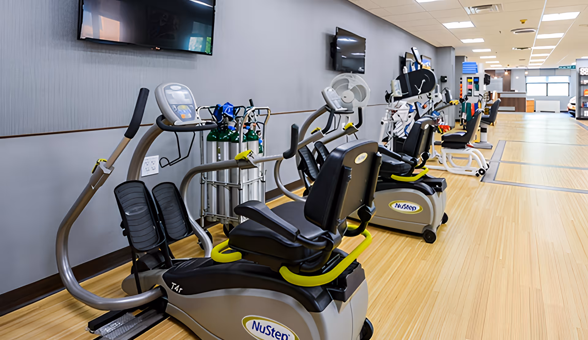 A rehabilitation center gym area with multiple exercise machines including NuStep recumbent cross trainers, oxygen tanks, and mounted televisions on the wall. The room has light wood flooring and gray walls with bright overhead lighting.