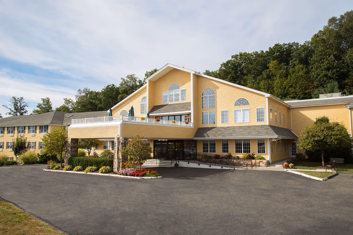Front exterior of a large yellow senior living building with a covered entrance, balcony, and landscaped driveway.