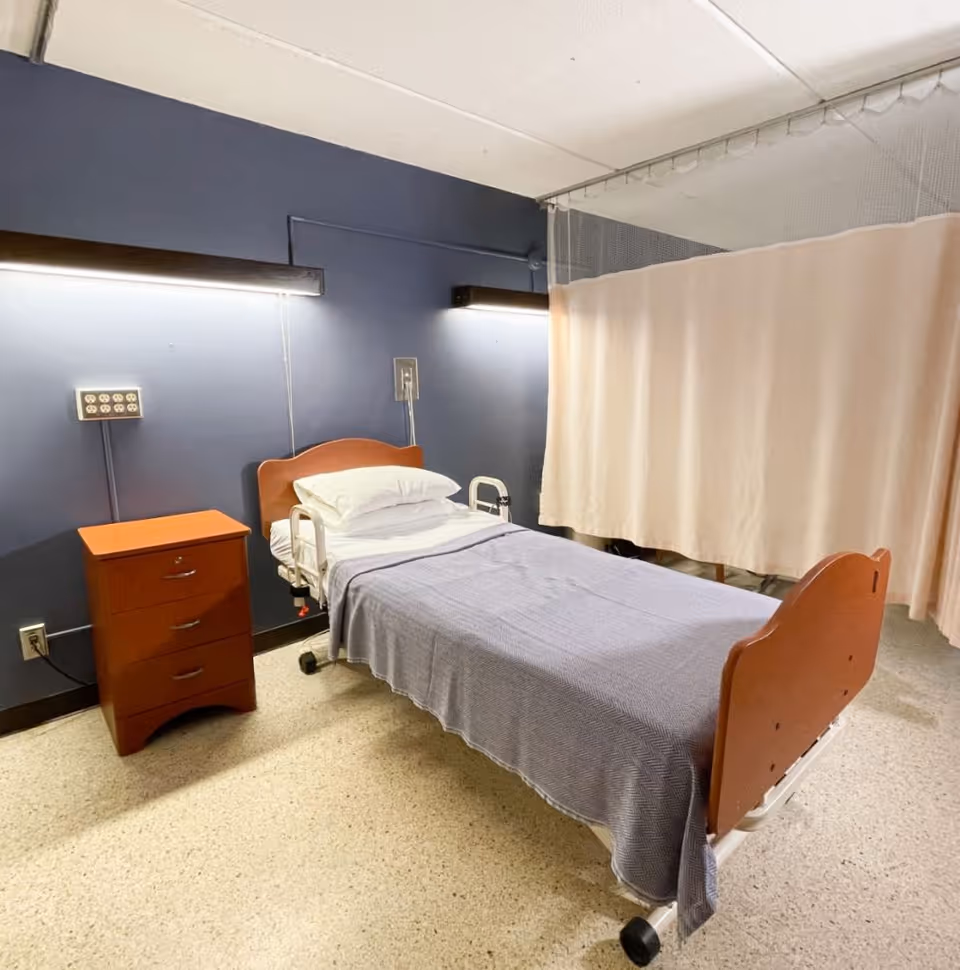 A single hospital-style bed with white pillows and a blue blanket beside a wooden nightstand and a beige privacy curtain in a clinical room.