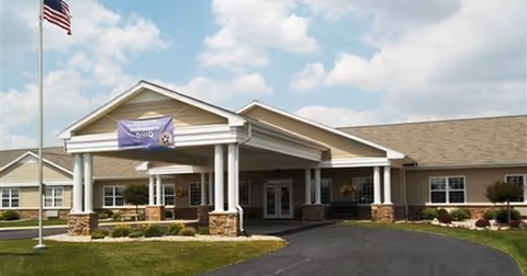 Front entrance of a single-story senior living building with a covered porte-cochere, an American flag, and landscaped lawn under a partly cloudy sky.