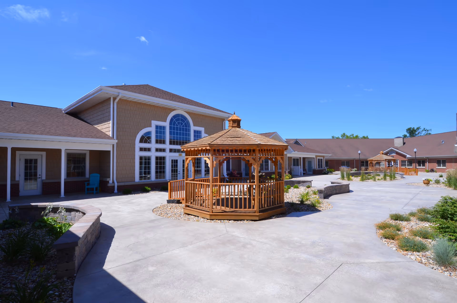 Outdoor courtyard area of Snyder Village senior living facility featuring two wooden gazebos, paved walkways, landscaped garden beds, and buildings with large windows under a clear blue sky.