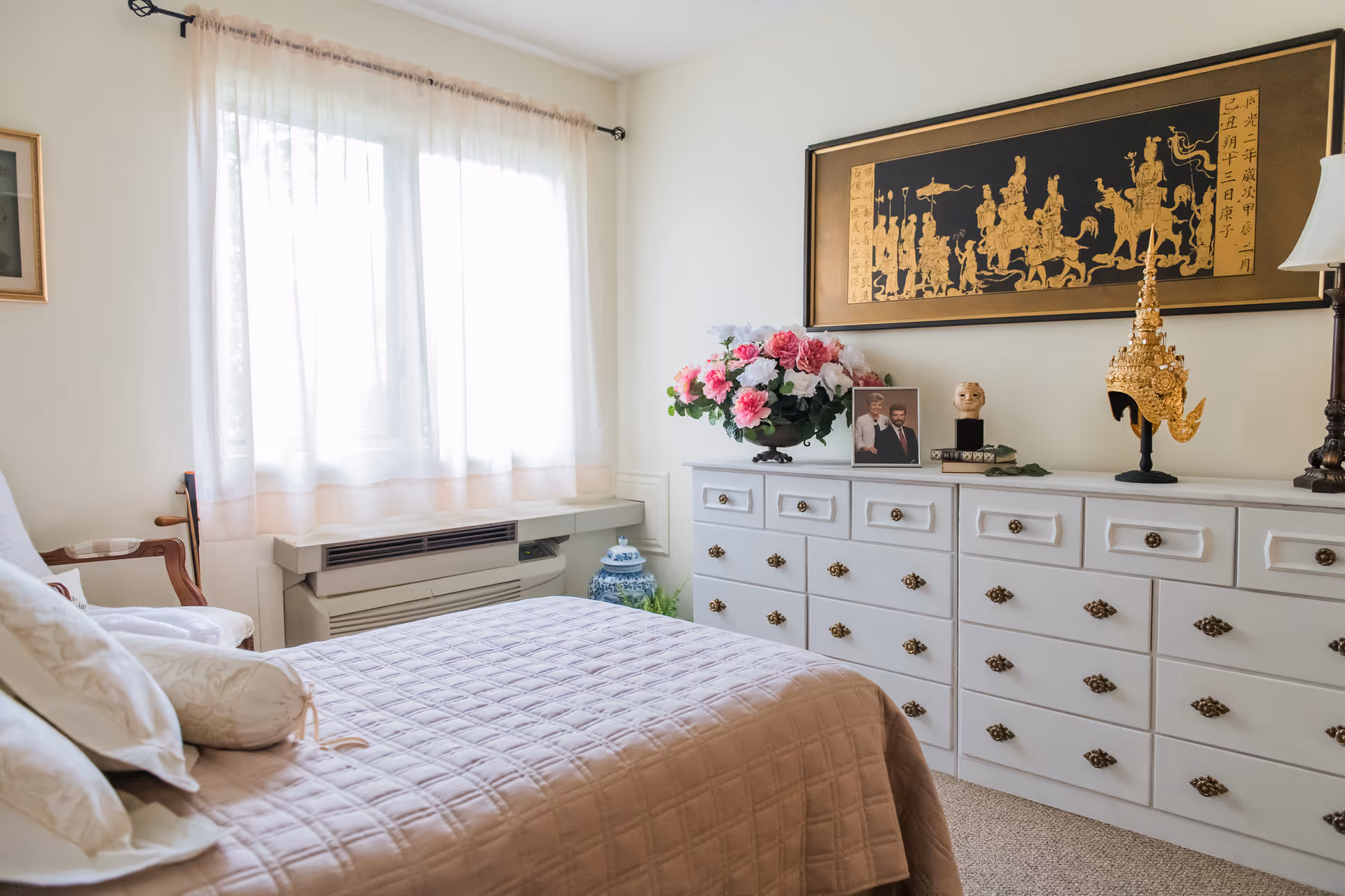A cozy bedroom with a bed covered in a light pink quilt and several pillows. Next to the bed is a wooden chair with a cushion. A large window with sheer white curtains allows natural light to fill the room. Along one wall is a white dresser with multiple drawers, adorned with a vase of pink and white flowers, a framed photo, a decorative mask, and a golden ornamental piece. Above the dresser hangs a large framed artwork with gold figures on a black background.