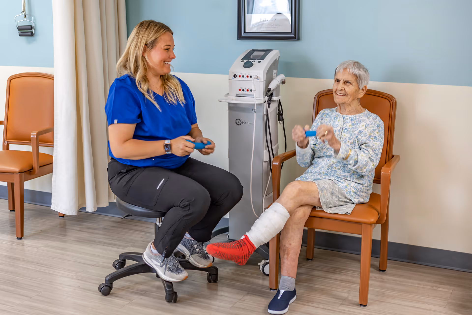A healthcare worker in blue scrubs sits on a rolling stool smiling and engaging with an elderly woman who has a bandaged leg and is seated in a chair. Both are holding small blue exercise weights in a clinical room with light blue and beige walls and medical equipment in the background.