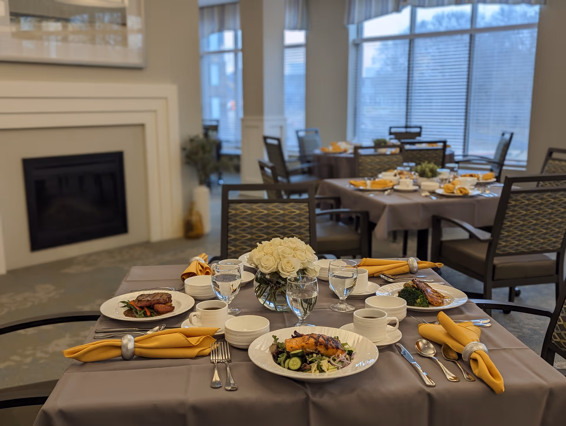 A dining room in a senior living facility with tables set for a meal. The tables have gray tablecloths, yellow napkins with silver napkin rings, plates with food including salad and meat, cups, glasses of water, and a centerpiece of white flowers. The room has large windows with blinds and a fireplace on one side.