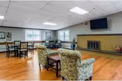 A senior living common lounge with armchairs and small tables facing a brick fireplace and wall-mounted TV, with windows along the back wall.