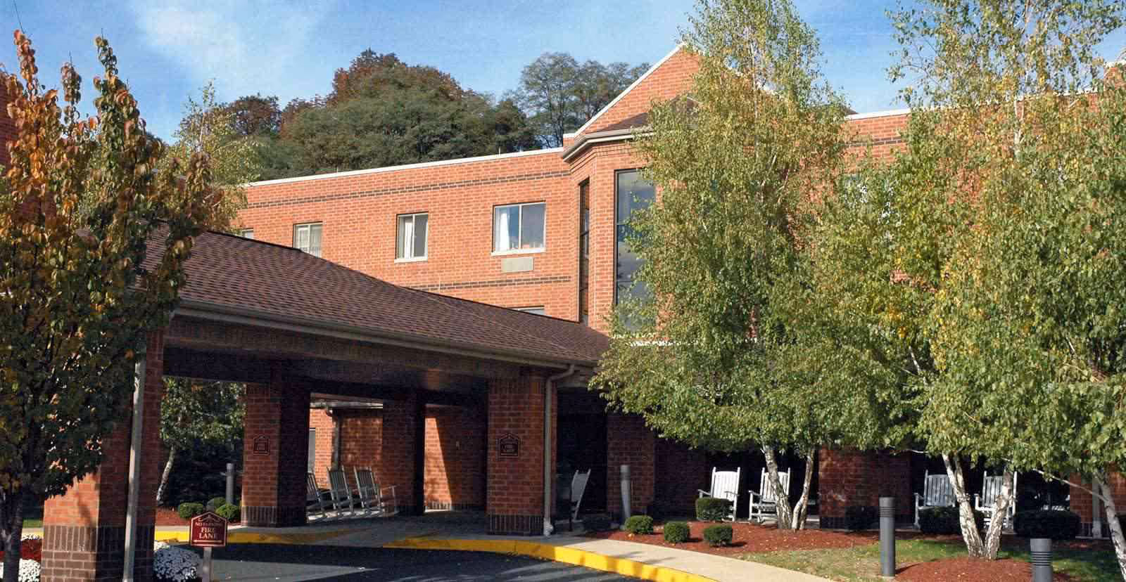 Front entrance of a brick senior living building featuring a covered porte-cochère, rocking chairs and landscaped trees.