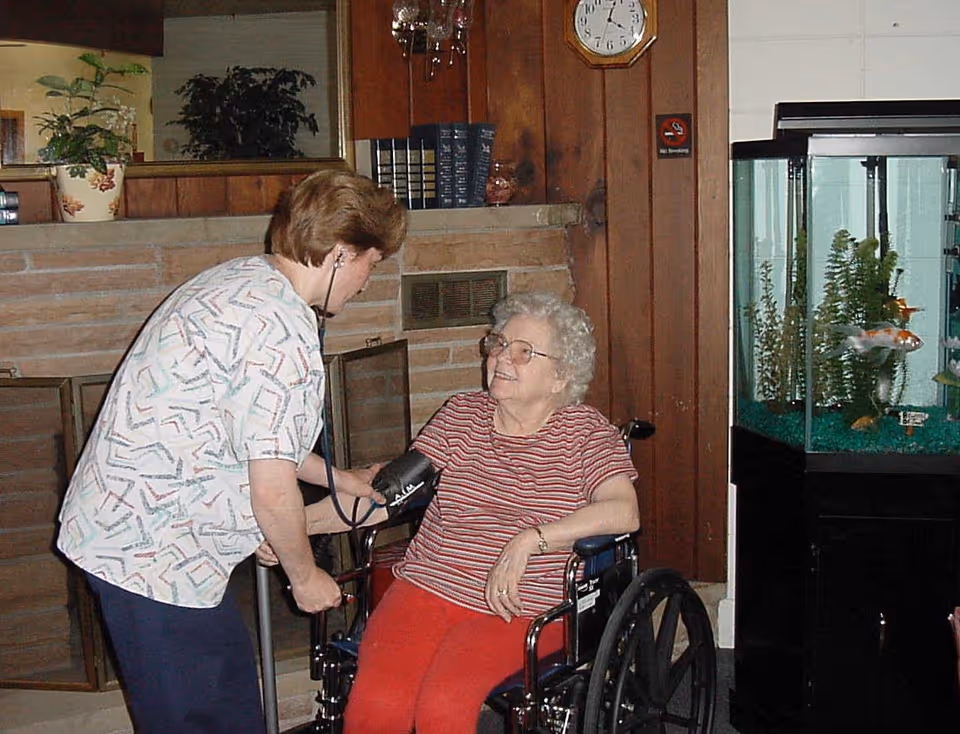 A caregiver is checking the blood pressure of an elderly woman seated in a wheelchair inside a room with wood-paneled walls, a fish tank, and a clock on the wall.