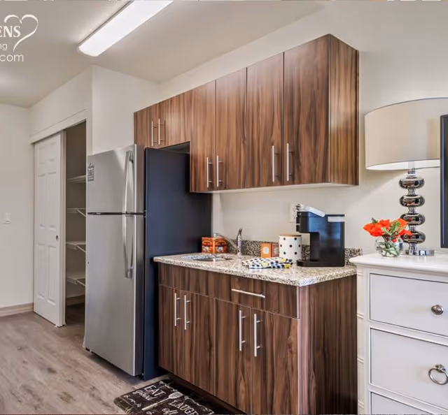 Small kitchenette with stainless steel refrigerator, wood cabinets, granite countertop, sink and a coffee maker with decorative items.