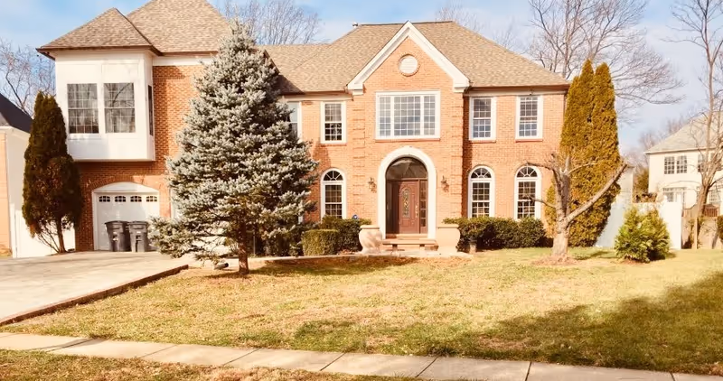 Front exterior view of a large two-story brick house with multiple windows, a central arched doorway, a driveway with trash bins, and a lawn with trees and shrubs.