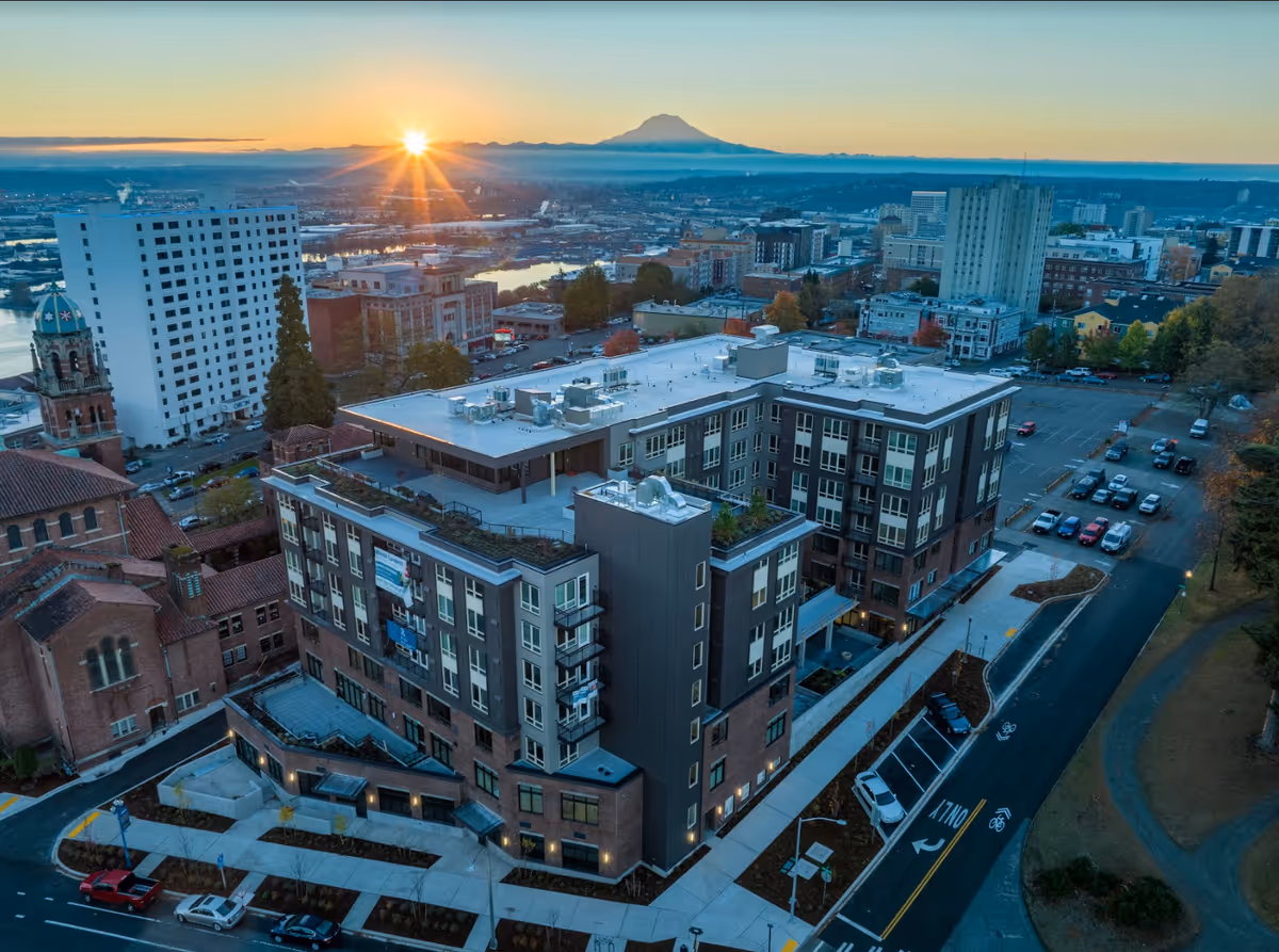 Aerial view of Merrill Gardens at Wright Park building at sunset, with the sun setting behind distant mountains and a cityscape in the background. The multi-story building is surrounded by streets, parked cars, and nearby structures including a church with a dome.