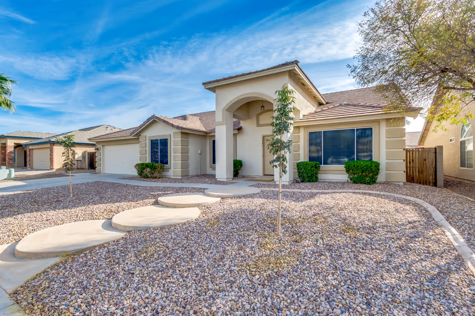 Single-story residential building with a beige exterior, a tiled roof, and an arched entrance. The front yard is landscaped with gravel, a few small trees, and bushes near the windows. A concrete pathway with circular stepping stones leads to the front door. The sky is clear with some wispy clouds.