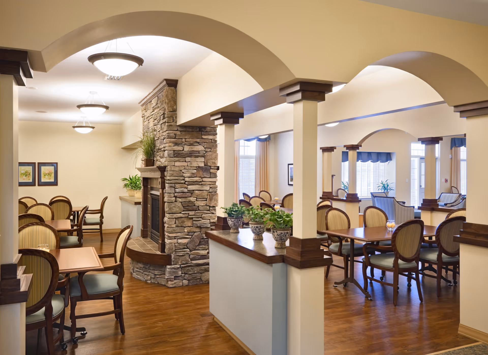 Interior view of a senior living facility dining area with wooden tables and chairs arranged neatly. The room features a stone fireplace, arched doorways, potted plants on a half wall, and warm lighting from ceiling fixtures. Large windows with curtains allow natural light to brighten the space.