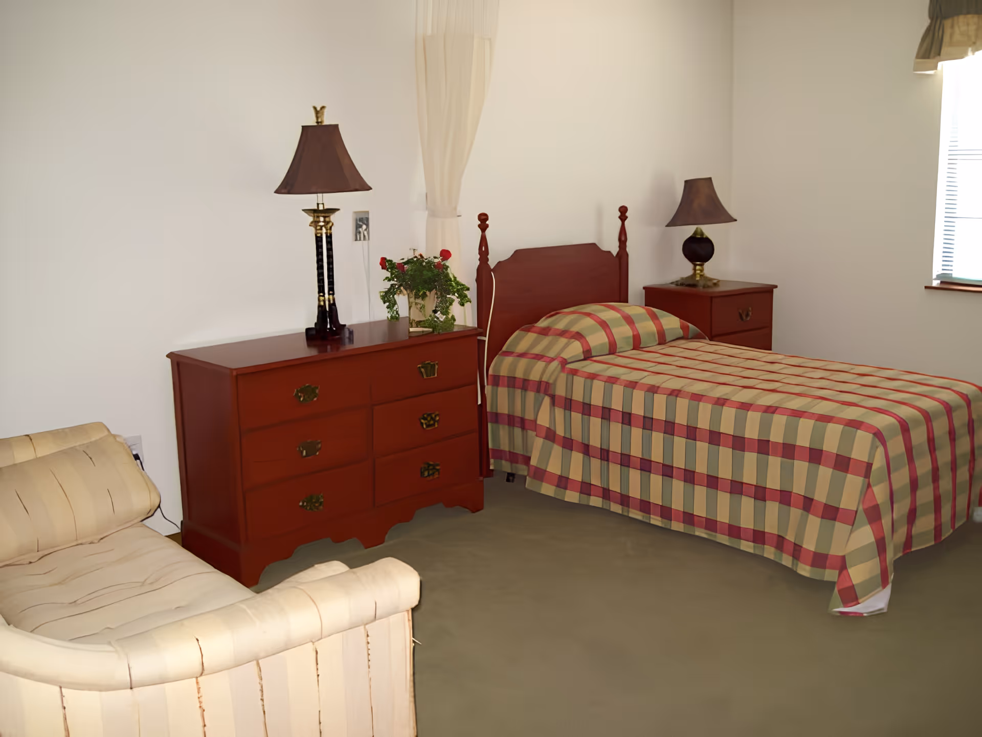 A bedroom with a single bed covered in a plaid bedspread in shades of beige, red, and green. There is a wooden headboard and matching wooden nightstand with a lamp on it. Next to the bed is a wooden dresser with six drawers, a lamp, and a potted plant with red flowers. A beige upholstered armchair is positioned near the dresser. The room has light-colored walls and a window with blinds partially open.