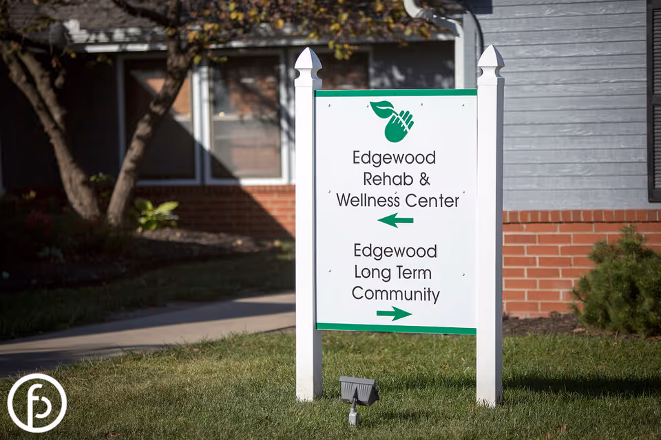 A white signpost with green accents outside a building, displaying directions for Edgewood Rehab & Wellness Center to the left and Edgewood Long Term Community to the right. The background includes a brick and gray siding building with a window and some greenery.