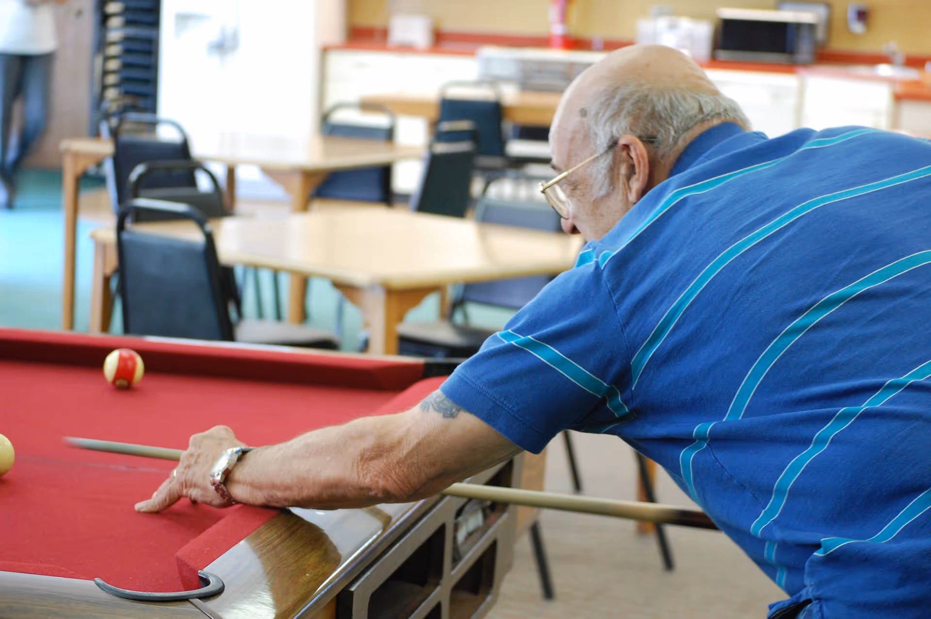 An elderly man wearing a blue striped shirt is playing pool on a red felt pool table in a common area with tables and chairs in the background.