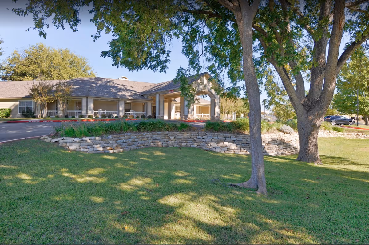 Exterior view of Sodalis Temple facility showing a single-story building with a covered entrance, surrounded by well-maintained green lawn, trees, and a stone retaining wall under a clear blue sky.