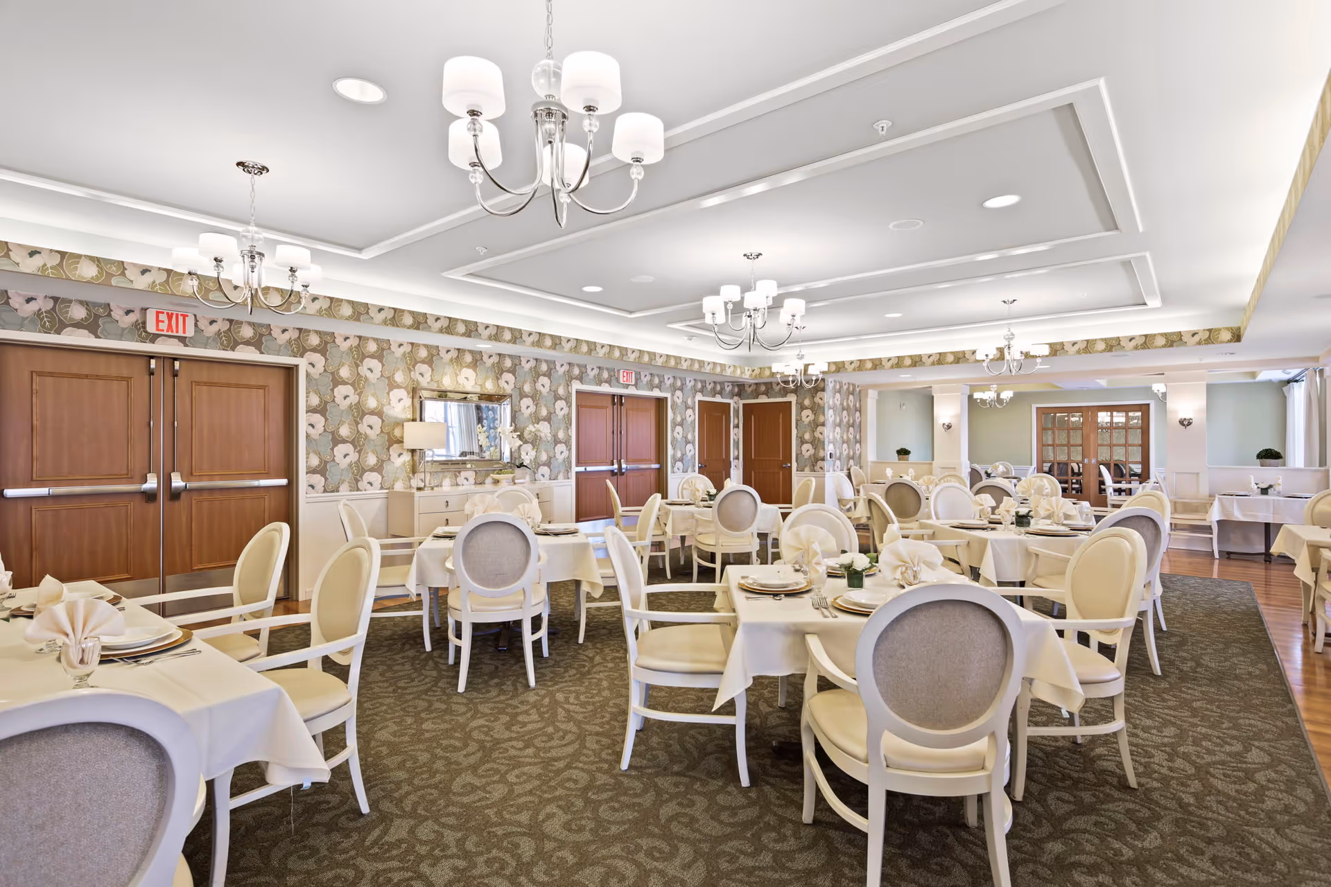 A spacious, elegantly decorated dining room with multiple round tables covered with white tablecloths and set with plates, napkins, and glassware. The room features floral wallpaper, several chandeliers, wooden double doors, and a carpeted floor with a patterned design.