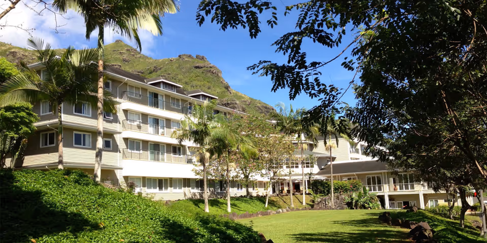 Exterior view of a multi-story senior living facility surrounded by lush greenery, palm trees, and a well-maintained lawn with a mountainous backdrop under a clear blue sky.