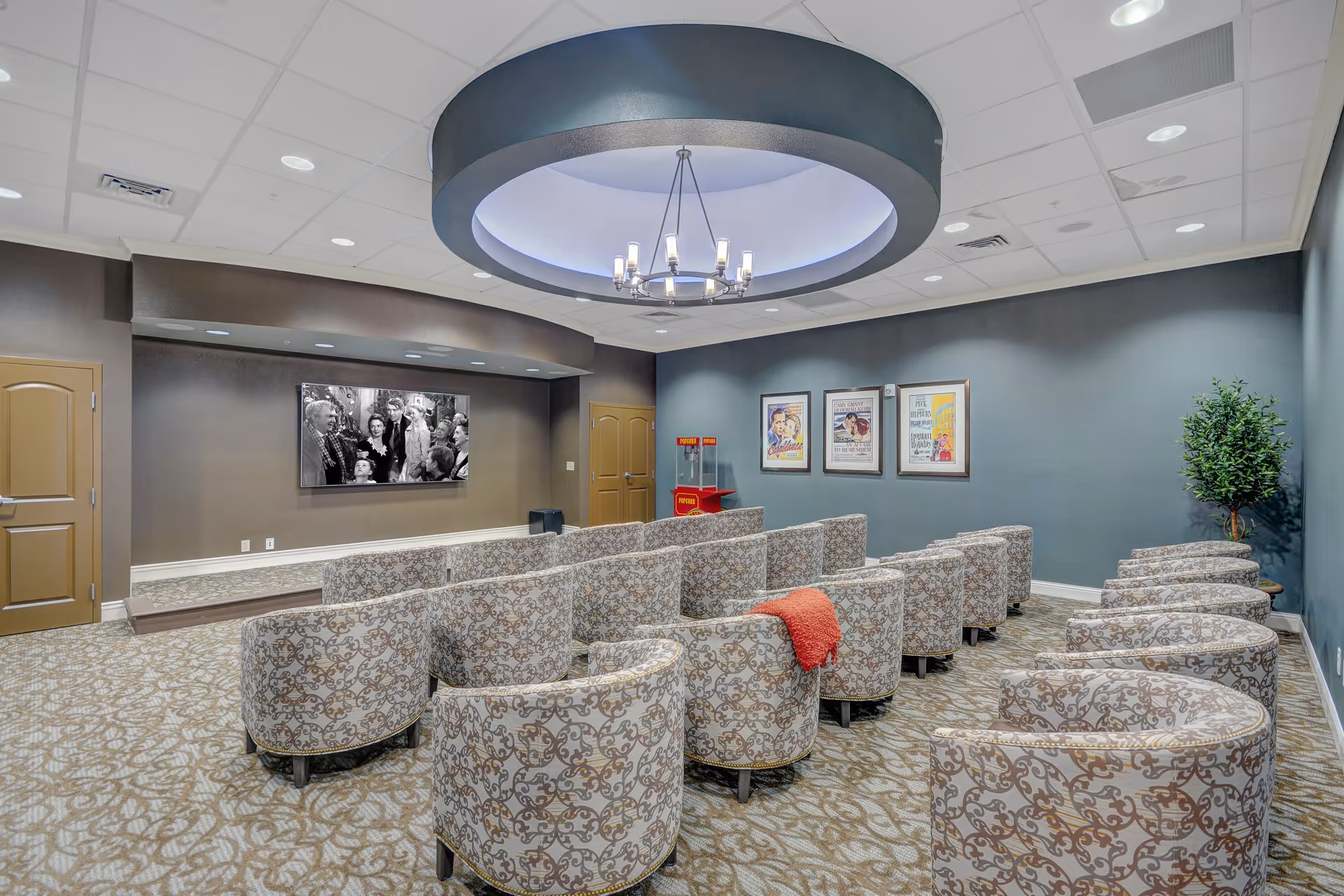 A cozy movie theater room with patterned armchairs arranged in rows facing a large screen showing a black and white movie. The room has a circular ceiling light fixture, three framed vintage movie posters on the wall, a popcorn machine in the corner, and a potted plant. The carpet and walls have a neutral color scheme.
