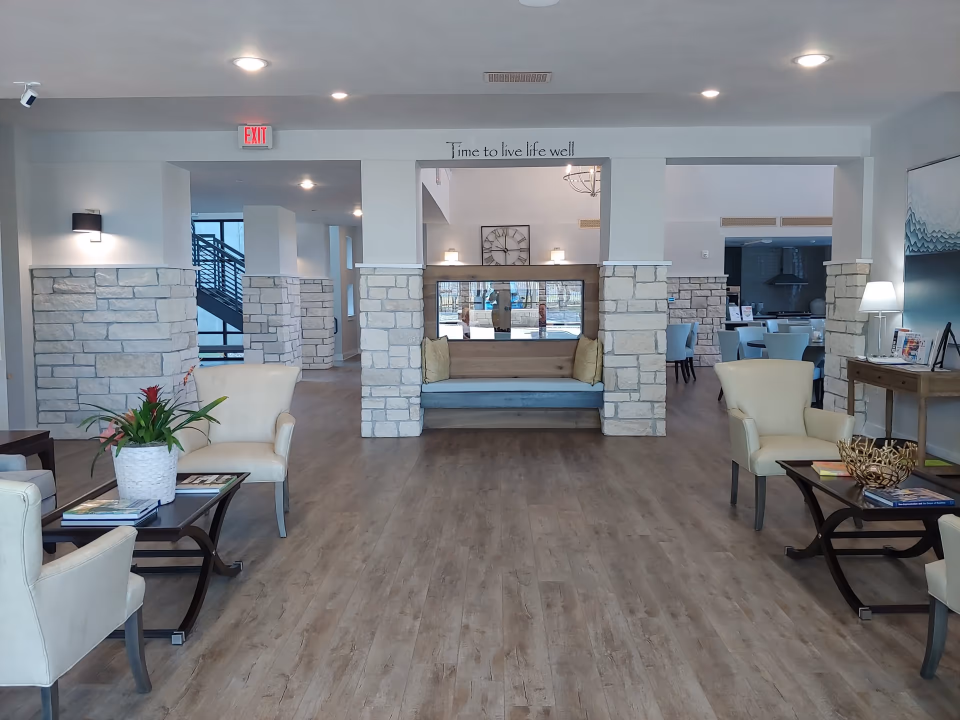 A spacious and well-lit senior living facility common area with light-colored walls and stone pillars. The room features comfortable cream-colored armchairs arranged around dark wood coffee tables with books and decorative items. A built-in bench with cushions is centered between two stone pillars, and a large clock hangs on the wall above it. The floor is wood, and there is a visible dining area with tables and chairs in the background. The ceiling has recessed lighting, and a sign above the bench reads 'Time to live life well'.