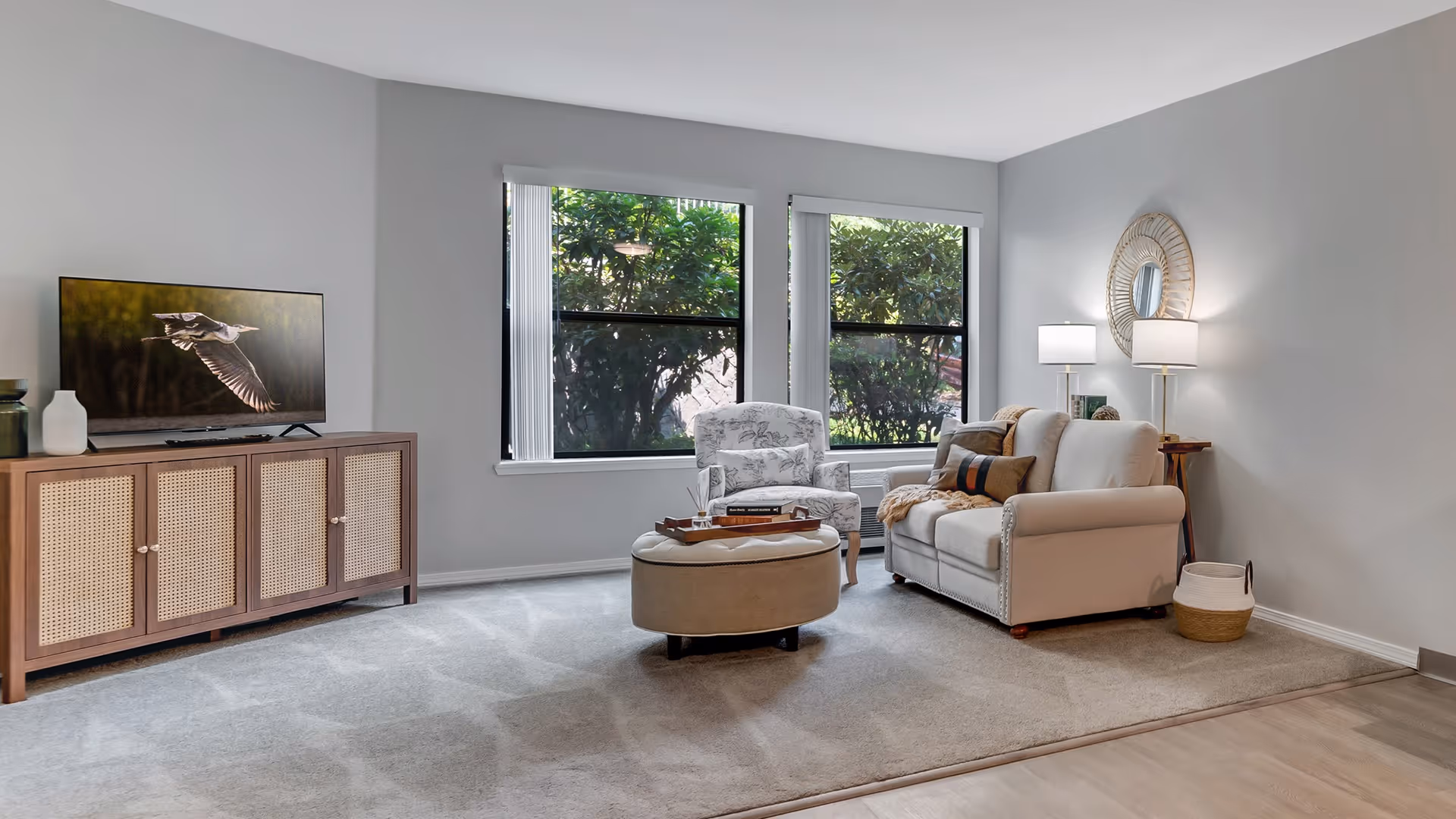 A cozy living room with a beige loveseat, a patterned armchair, and a round ottoman in the center. There is a wooden cabinet with a flat-screen TV displaying a bird in flight. Two large windows with vertical blinds let in natural light, and a round decorative mirror hangs on the wall above a side table with two lamps. The floor is carpeted with a light gray rug, and there is a basket on the floor next to the loveseat.