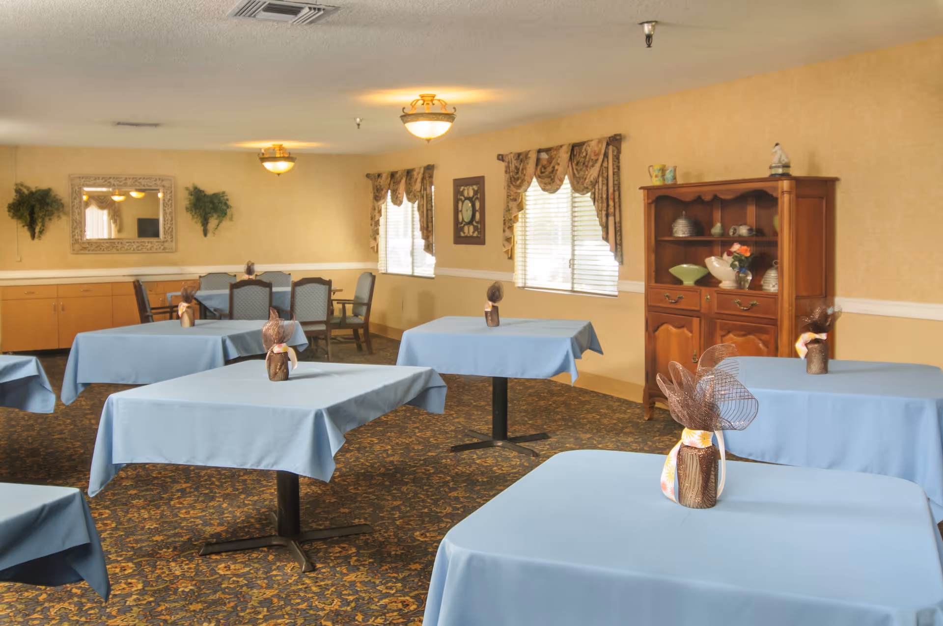 A dining room with several square tables covered in light blue tablecloths, each decorated with a small centerpiece. The room has patterned carpet, beige walls with white trim, two windows with floral curtains, a wooden cabinet with decorative items, and a mirror on one wall.