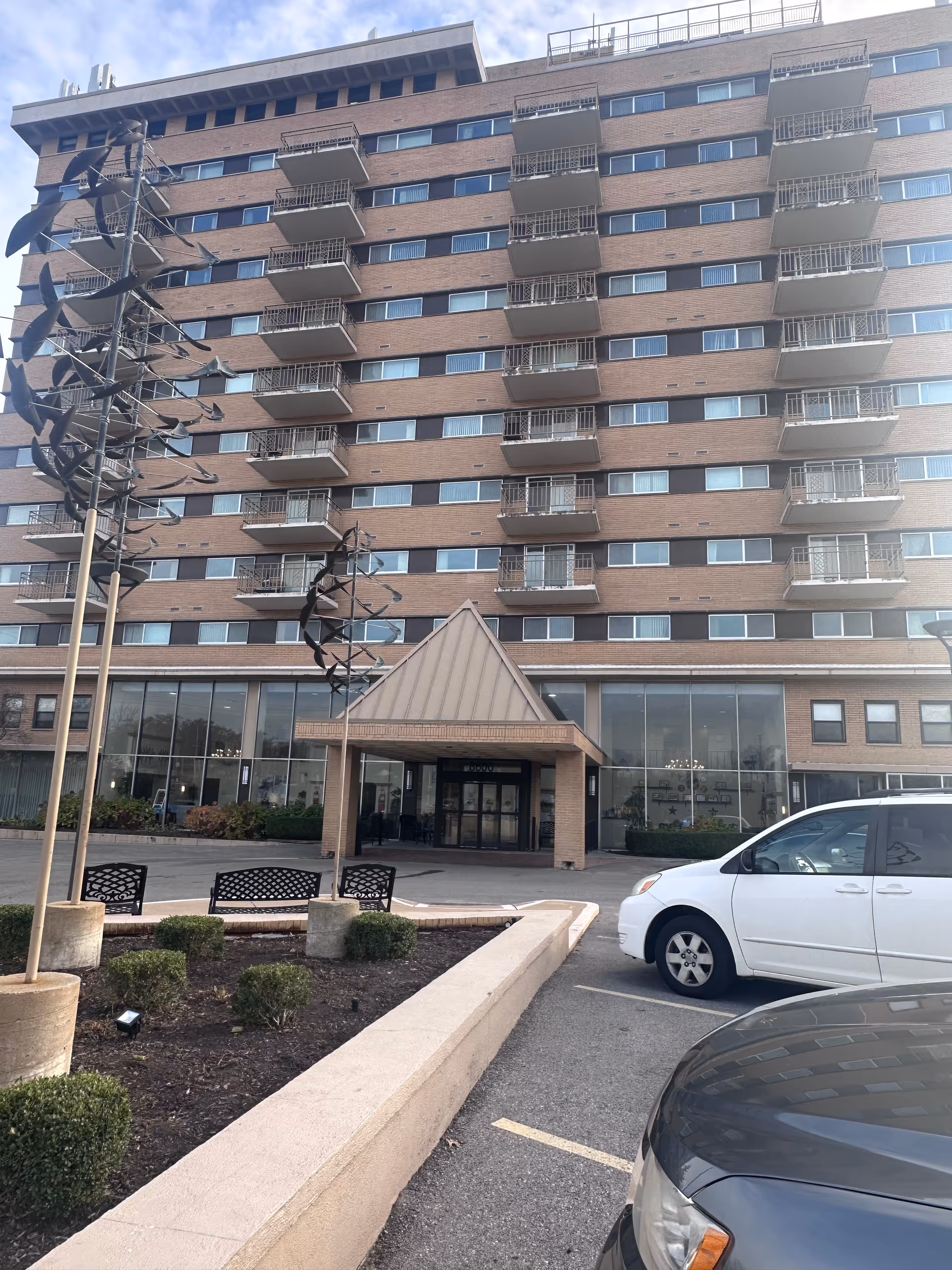 Exterior view of a multi-story brick building with balconies, a covered entrance, and large windows on the ground floor. There are two cars parked in front and some landscaped bushes and benches near the entrance.
