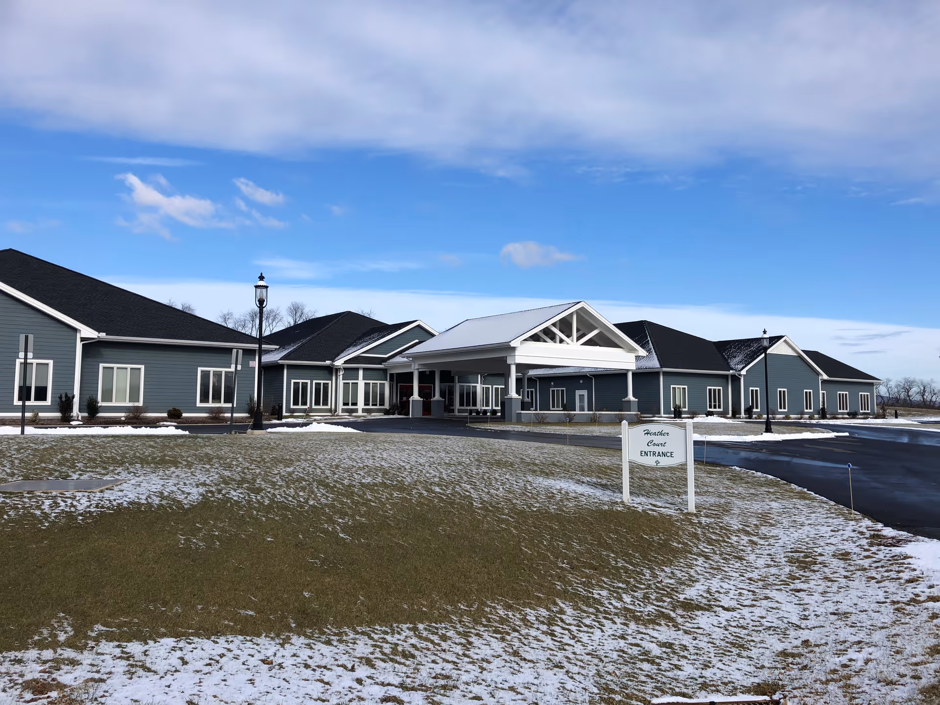 Exterior view of Heather Court at Nottingham Village, a single-story building with blue-gray siding and a white covered entrance. The surrounding lawn is partially covered with snow under a partly cloudy blue sky.