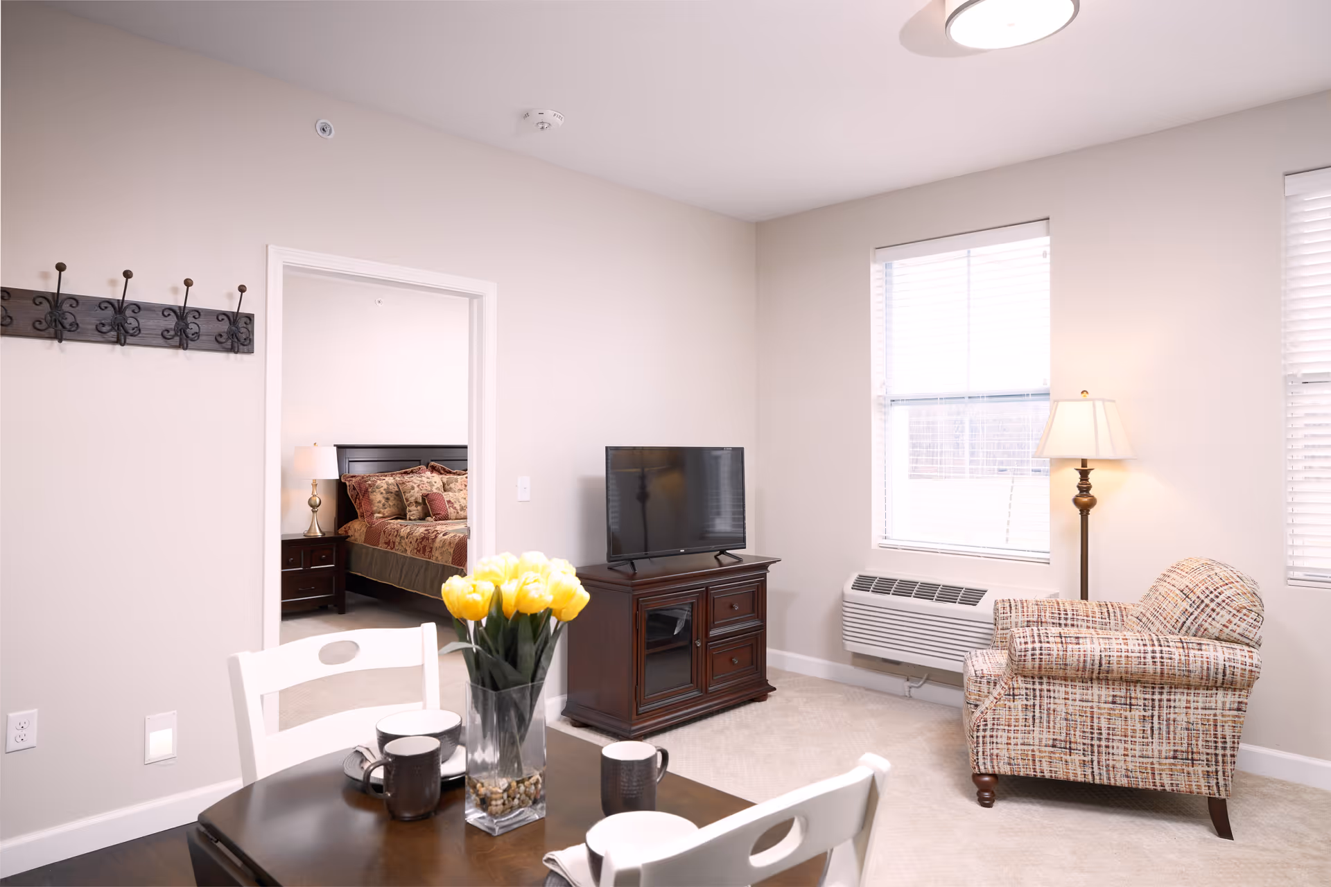 A bright living room area with a patterned armchair, a floor lamp, a TV on a wooden stand, and two windows with blinds. A dining table with white chairs and a vase of yellow tulips is in the foreground. Through an open doorway, a bedroom with a bed, nightstand, and lamp is visible.