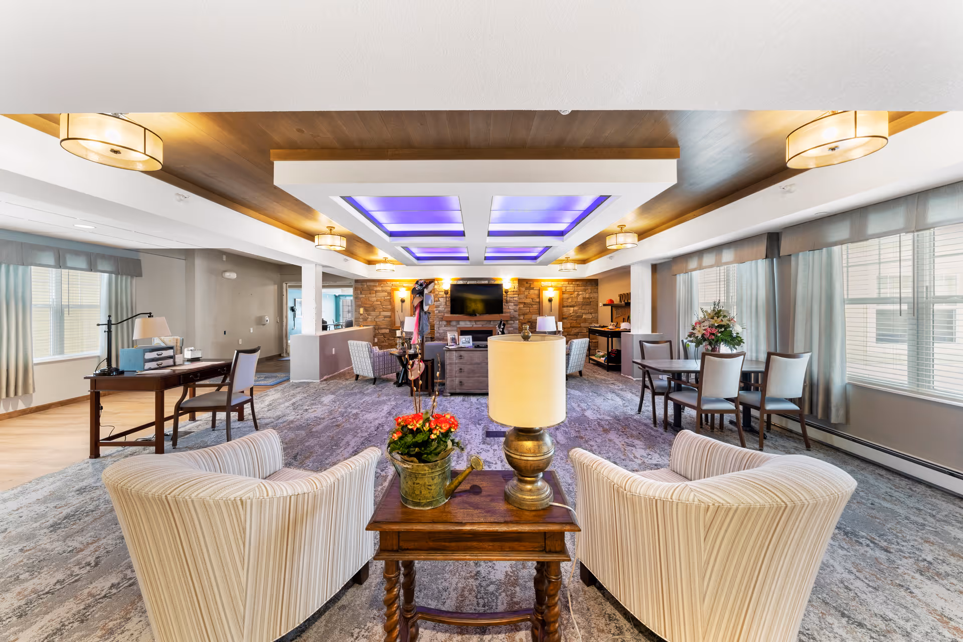 Bright, spacious senior living common room with upholstered armchairs and a side table in the foreground, dining table to the right, and a TV and seating area beneath decorative ceiling lights.