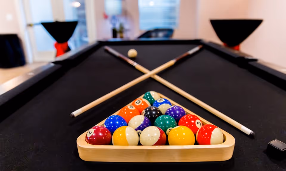 A black pool table with a wooden rack holding colorful billiard balls arranged in a triangle. Two pool cues are crossed and resting on the table behind the balls. The background shows a blurred interior space with high tables covered in black cloth and red sashes.