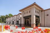 Exterior view of a beige stucco building with a tiled roof and an entrance canopy, surrounded by greenery and red flowers in the foreground under a clear blue sky.