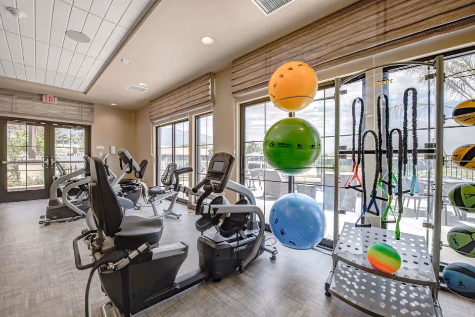 A bright fitness room with exercise equipment including stationary bikes and various fitness balls and resistance bands organized on racks. Large windows allow natural light to fill the space and offer a view of an outdoor patio area.