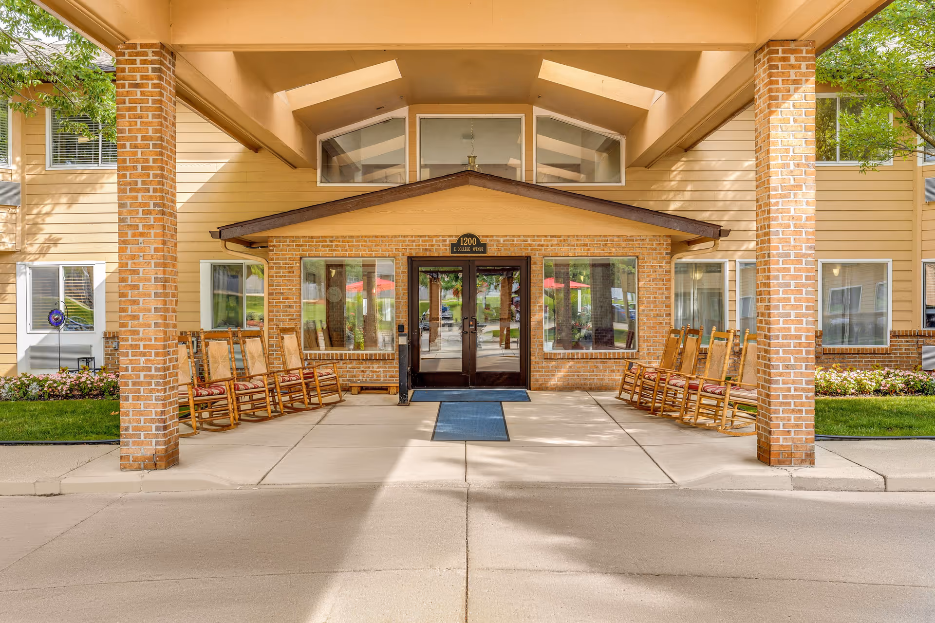 Entrance of Holiday Blair House facility featuring a covered driveway with brick pillars, wooden rocking chairs lined up on both sides, and double glass doors with windows on either side. The building exterior is beige with brick accents and has a sign above the door displaying the address 1200 E College Avenue.