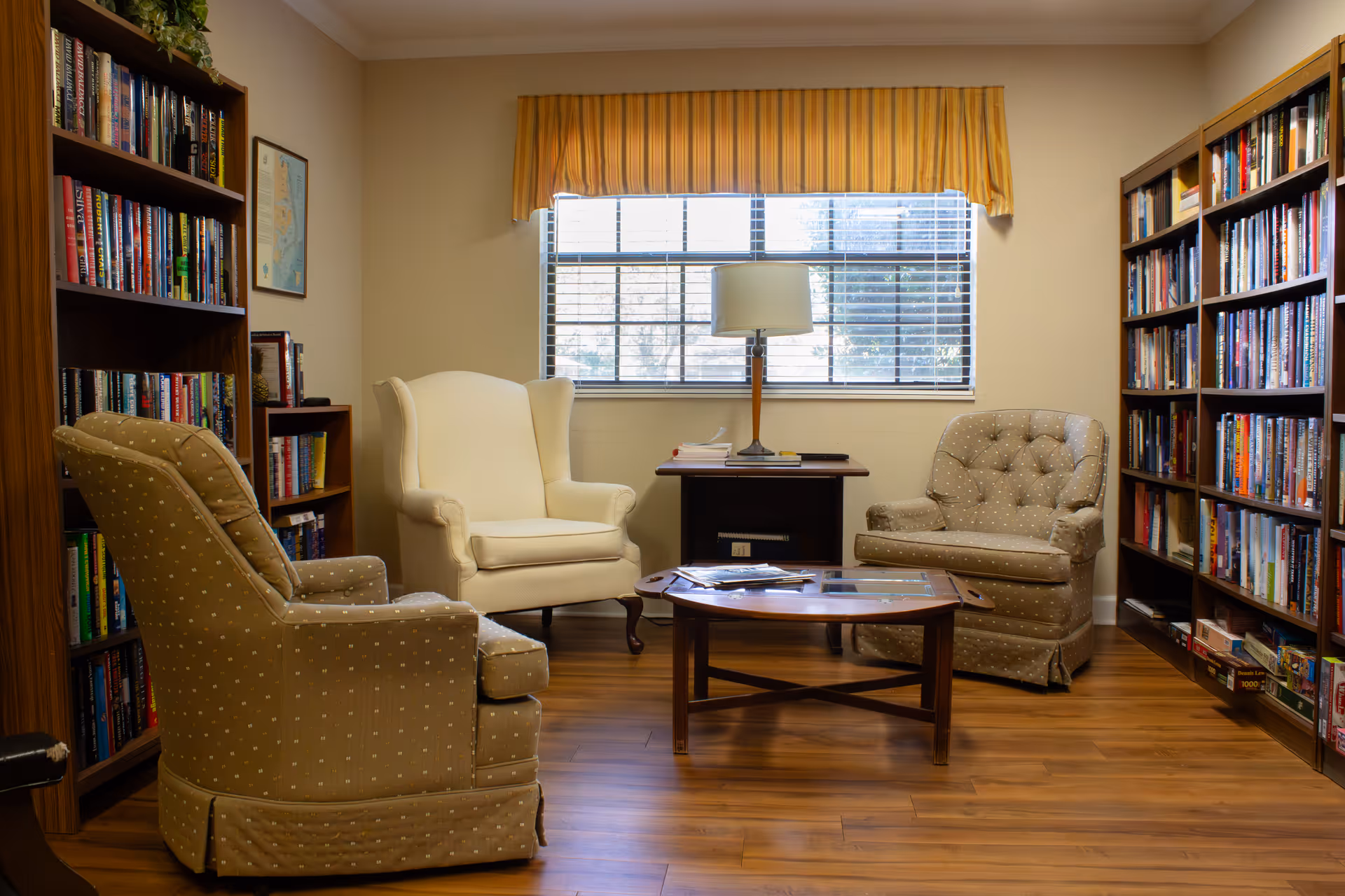 Cozy library-style sitting room with three armchairs around a wooden coffee table, bookshelves along the walls, and a window with a lamp.