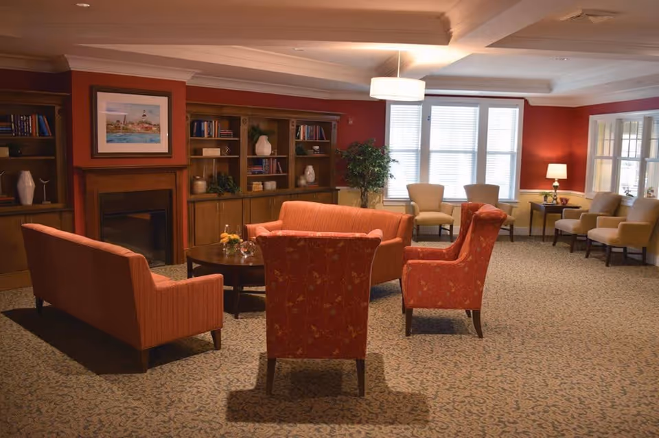 A cozy living room area with red and beige walls, featuring two red upholstered armchairs and a matching sofa arranged around a round wooden coffee table. The room has a fireplace with a framed painting above it, built-in wooden bookshelves filled with books and decorative items, a large window with white blinds, several beige armchairs, a floor lamp, and a potted plant.