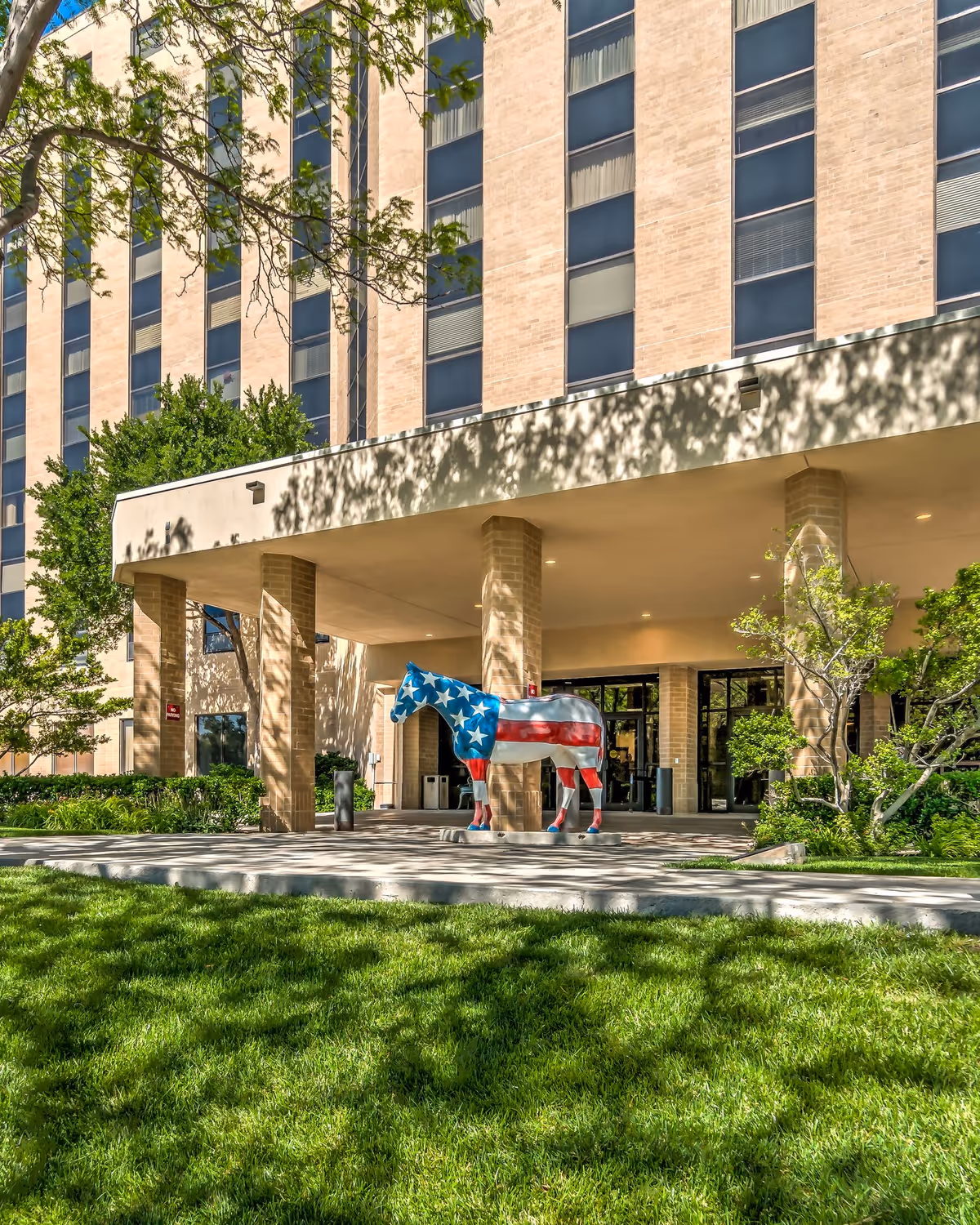 Entrance of a multi-story brick building with large windows, featuring a covered driveway supported by brick columns. In front of the entrance stands a horse sculpture painted with the American flag design. Green grass and trees surround the area, casting shadows on the ground.
